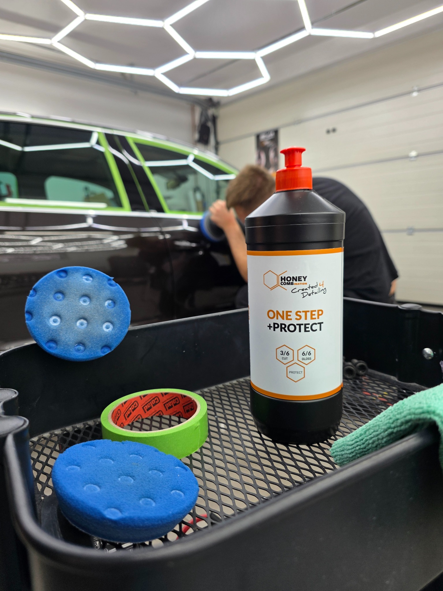 Polerowanie auta pastą Honey Combination One Step + Protect. W tle auto z taśmą zabezpieczającą i polerka w użyciu. Na pierwszym planie akcesoria.