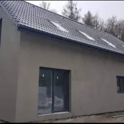 Dom jednorodzinny z szarą elewacją po tynkowaniu, z ciemnymi oknami i dachem pokrytym dachówką, widoczne ślady śniegu na dachu i wokół domu.