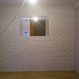 Ściana wyłożona dekoracyjną, białą cegłą w trakcie prac wykończeniowych w Kolnie, z widocznym otworem na okno lub drzwi i drewnianą podłogą.