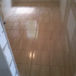 Beżowe płytki podłogowe ułożone w małej łazience, widoczne ślady fugowania na ścianie po lewej stronie.