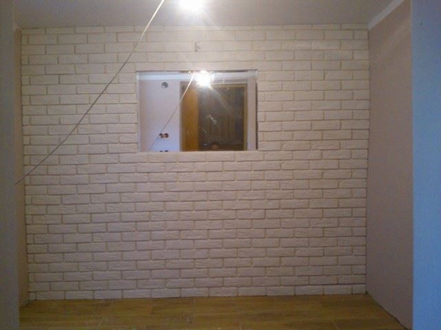 Ściana wyłożona dekoracyjną, białą cegłą w trakcie prac wykończeniowych w Kolnie, z widocznym otworem na okno lub drzwi i drewnianą podłogą.