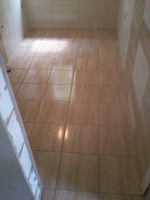 Beżowe płytki podłogowe ułożone w małej łazience, widoczne ślady fugowania na ścianie po lewej stronie.