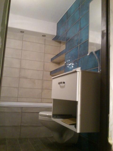 Wykończona łazienka z niebieskimi płytkami na ścianie, białą szafką pod umywalką i elementami armatury, widok z perspektywy wejścia.