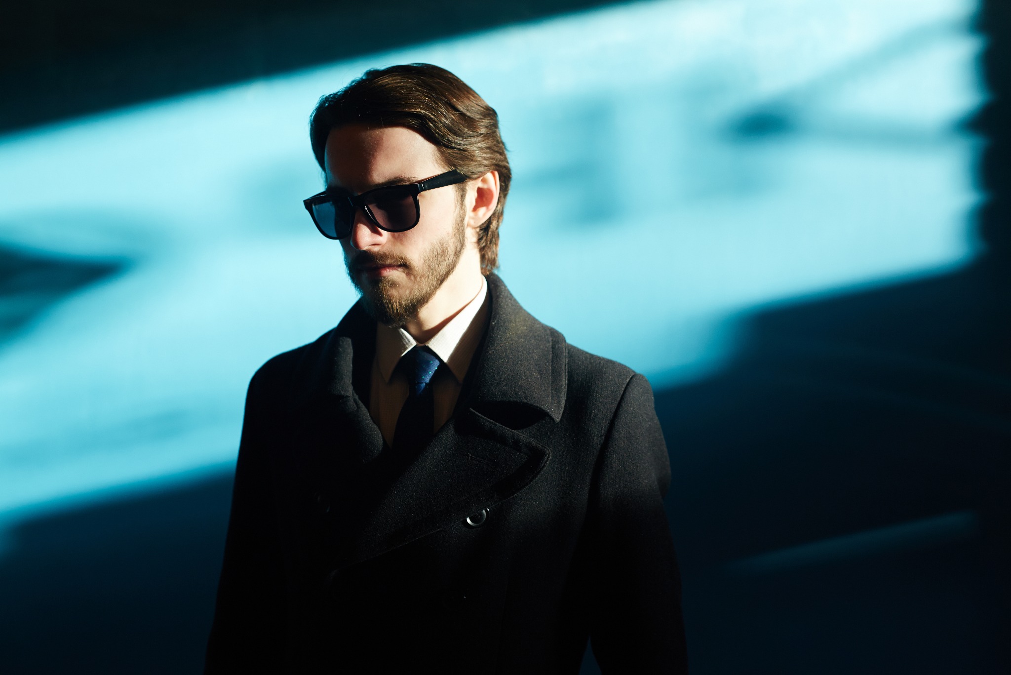 Mężczyzna w ciemnym płaszczu i okularach przeciwsłonecznych, oświetlony niebieskim światłem z góry.
