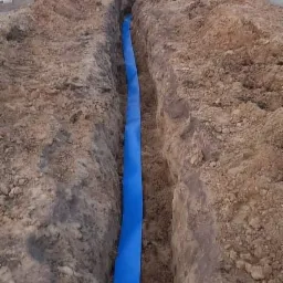 Wykop z niebieską rurą osłonową w ziemi, widok z góry, budynek w tle