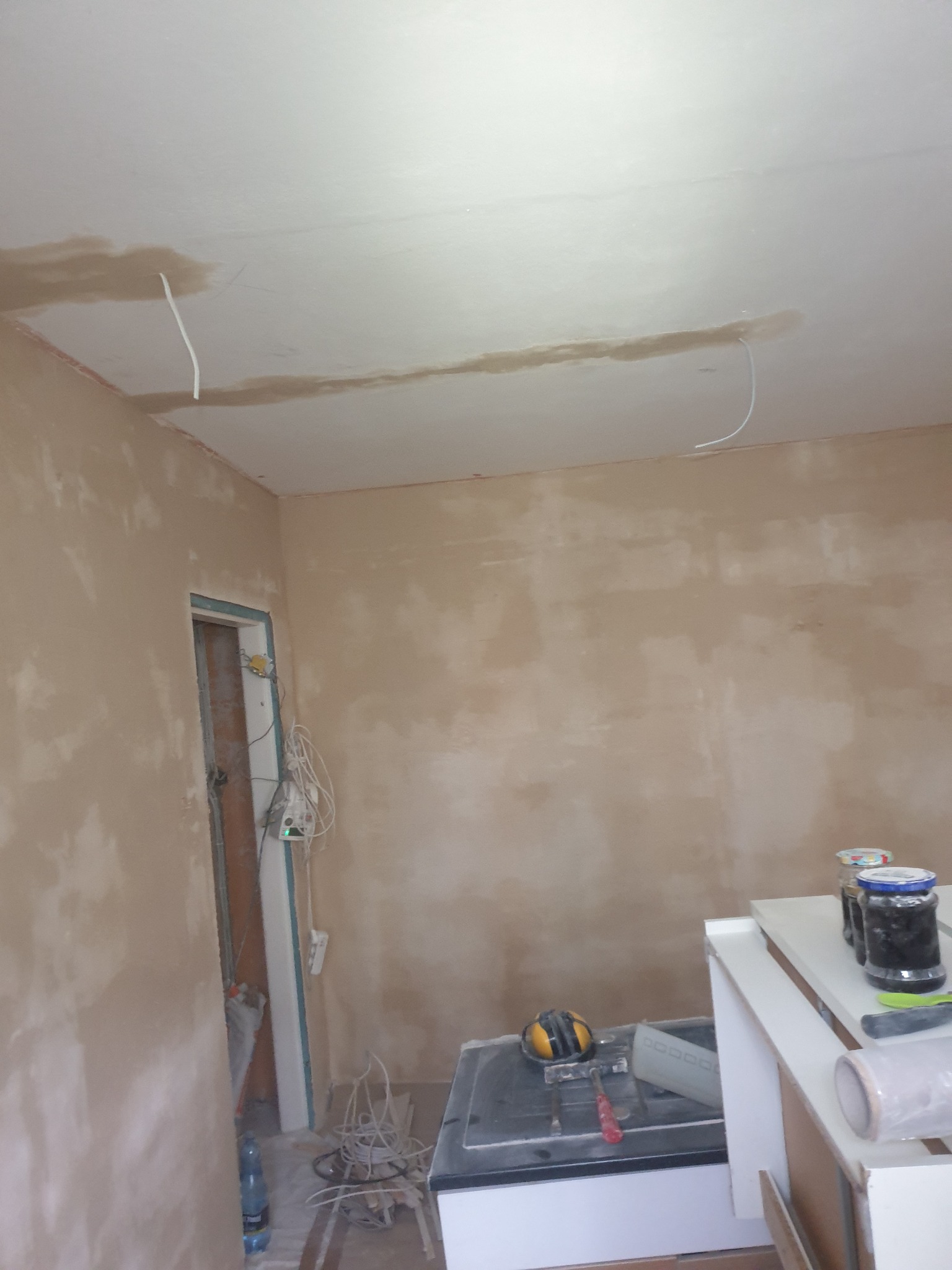 Pokój w trakcie remontu, ściany pokryte świeżym tynkiem, widoczne wystające przewody elektryczne z sufitu, przygotowane narzędzia i materiały budowlane.