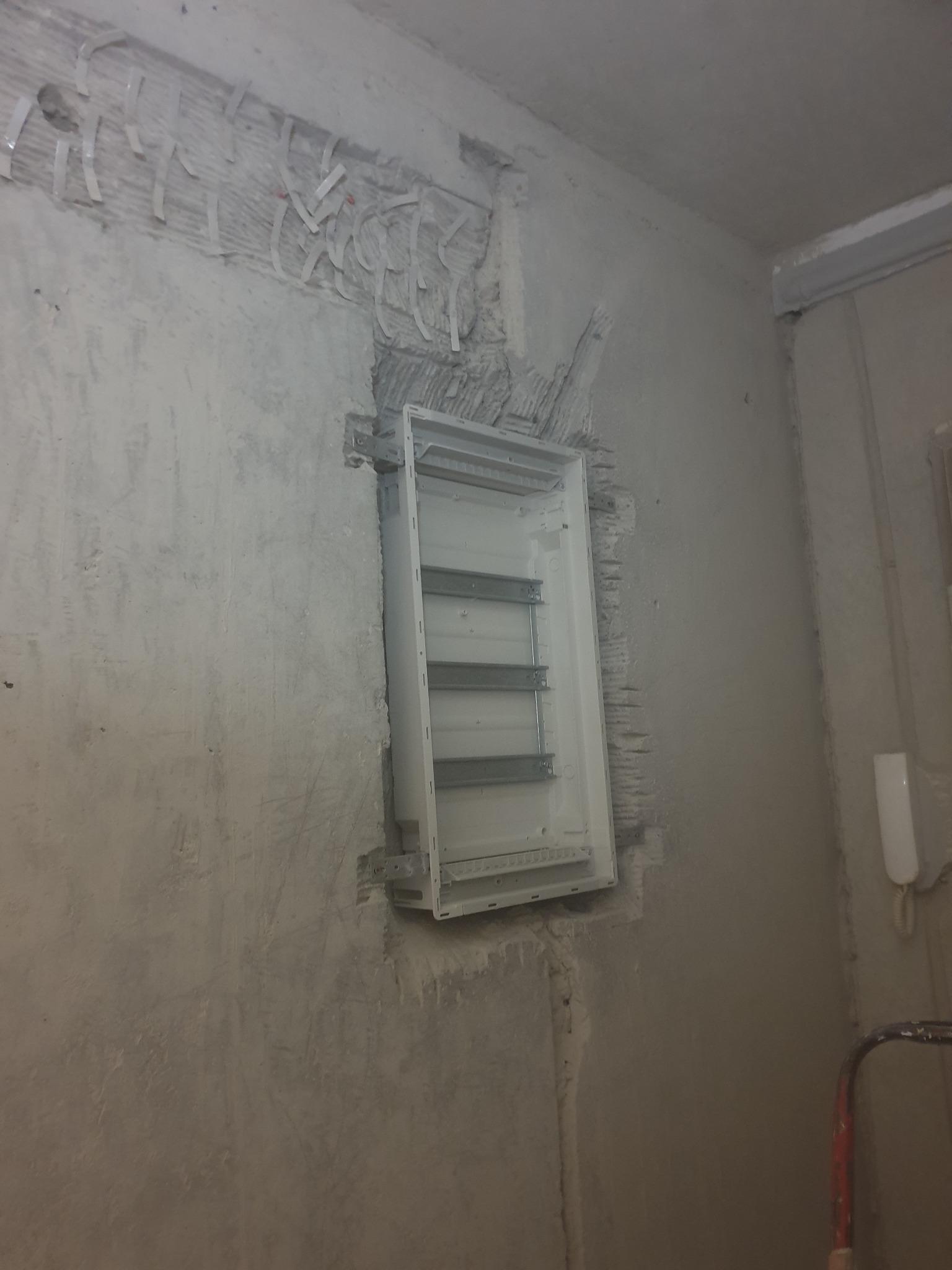 Wnęka w surowej ścianie przygotowana pod montaż skrzynki rozdzielczej z widocznymi przewodami zabezpieczonymi taśmą. Ściana wymaga wykończenia.