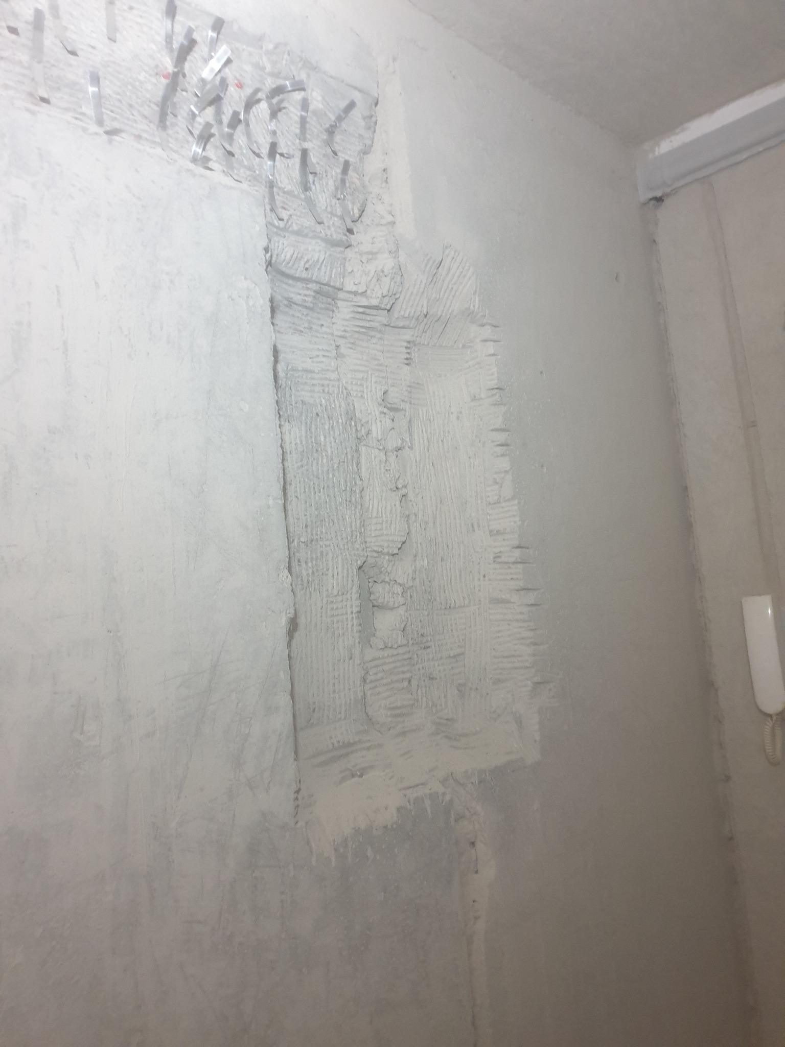 Ściana w trakcie remontu, z widocznymi przewodami elektrycznymi zabezpieczonymi klamrami i fragmentem ściany pokrytym grubą warstwą szpachli z charakterystyczną fakturą, przygotowywanej pod gładź.