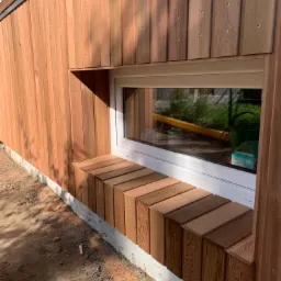 Drewniana elewacja budynku z oknem i parapetem wykonanym z drewnianych klocków, widoczne śruby mocujące deski elewacyjne.