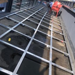 Merry Wood - Konstrukcja metalowa pod podłogę tarasu na dachu budynku, widoczne regulowane podstawki, w tle pracownicy i czerwone krzesło.