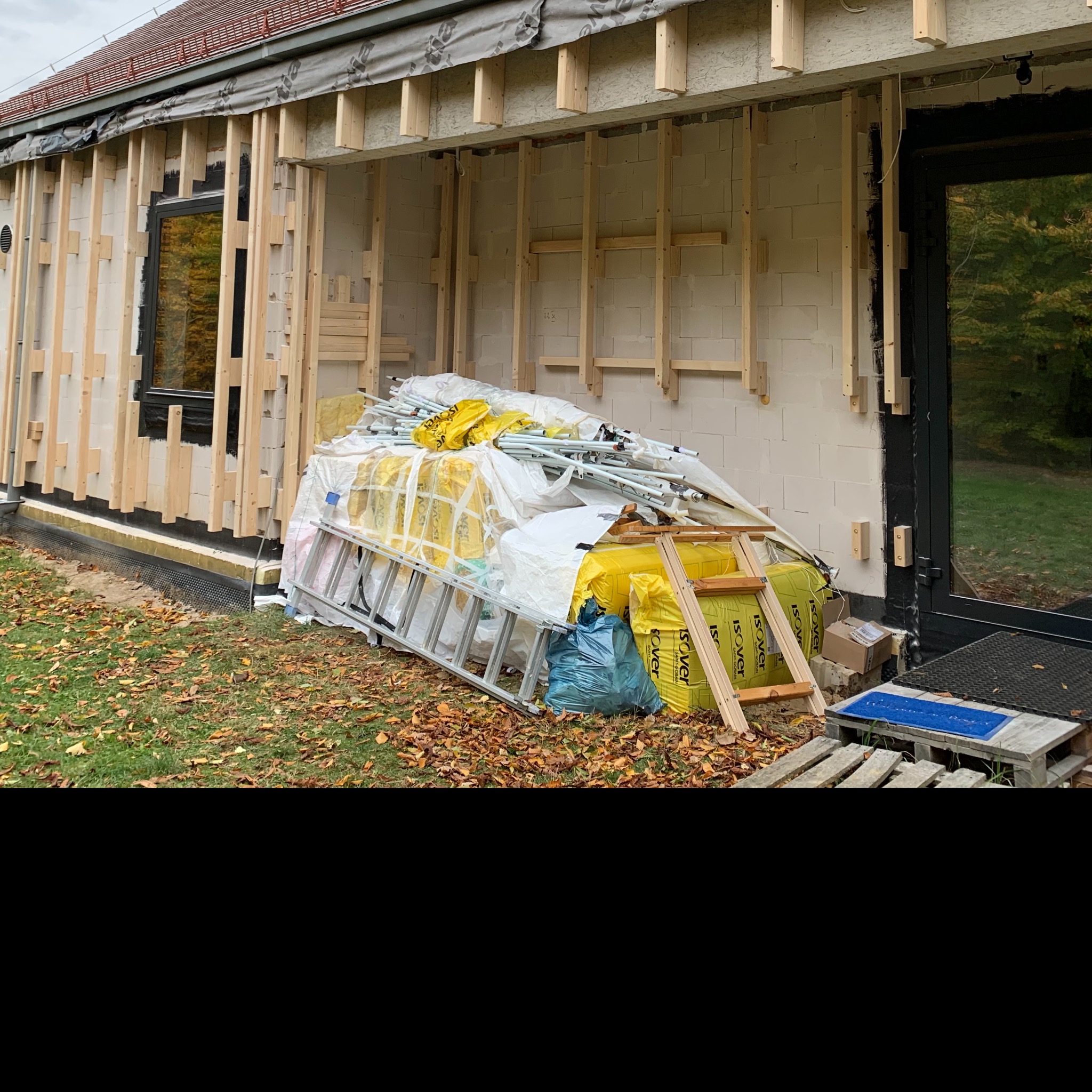 Budynek w trakcie ocieplania elewacji, widoczne drewniane ramy, materiały izolacyjne Isover, aluminiowa drabina i niebieski dywanik przed wejściem.