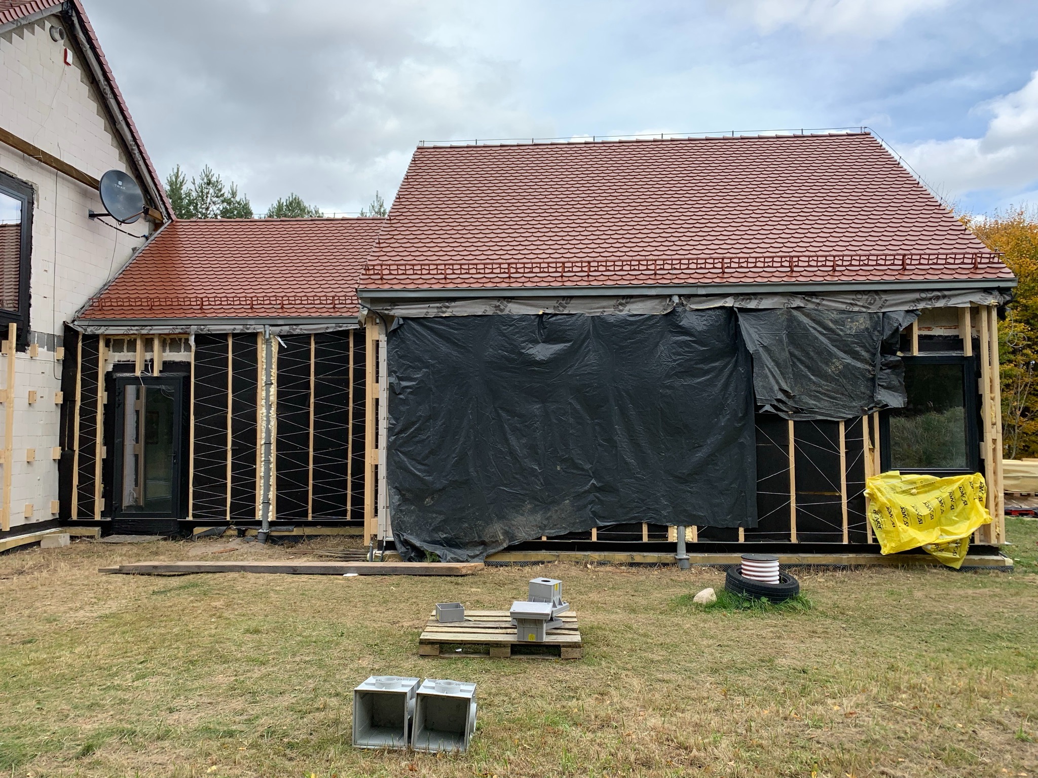 Budynek w trakcie ocieplania elewacji, widoczne drewniane ramy, czarna folia ochronna, czerwona dachówka, styropian w żółtej folii i elementy wentylacyjne na trawniku.