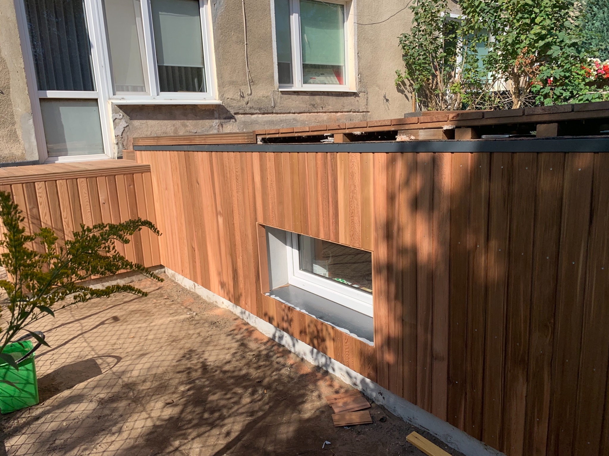 Nowa elewacja z drewnianych paneli pionowych, z małym oknem, częściowo zakrywająca starą elewację budynku.