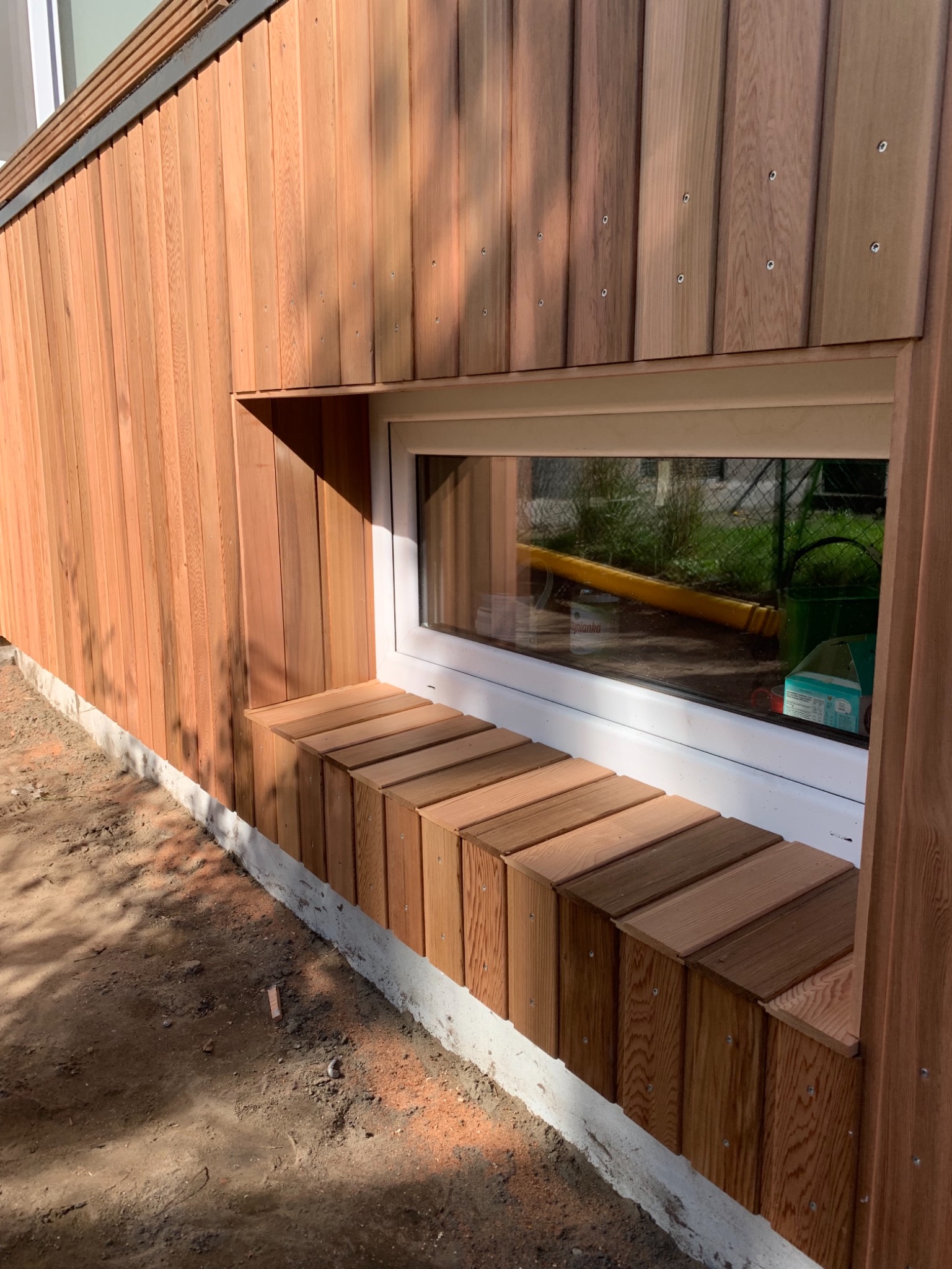 Drewniana elewacja budynku z oknem i parapetem wykonanym z drewnianych klocków, widoczne śruby mocujące deski elewacyjne.