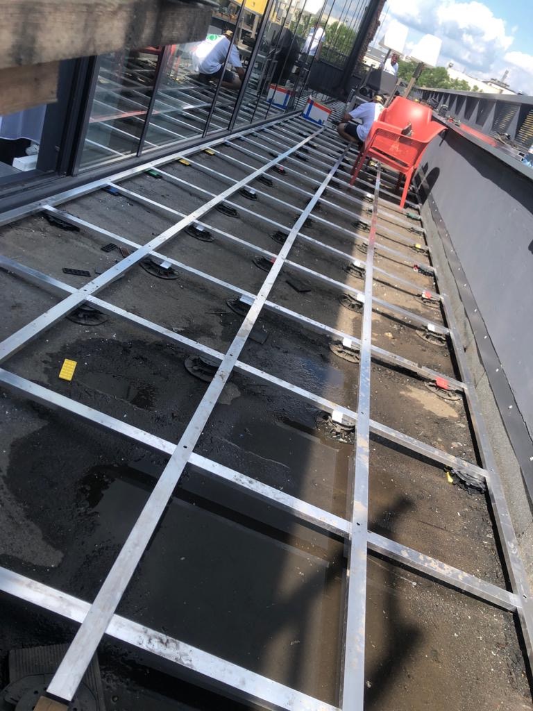 Konstrukcja metalowa pod podłogę tarasu na dachu budynku, widoczne regulowane podstawki, w tle pracownicy i czerwone krzesło.