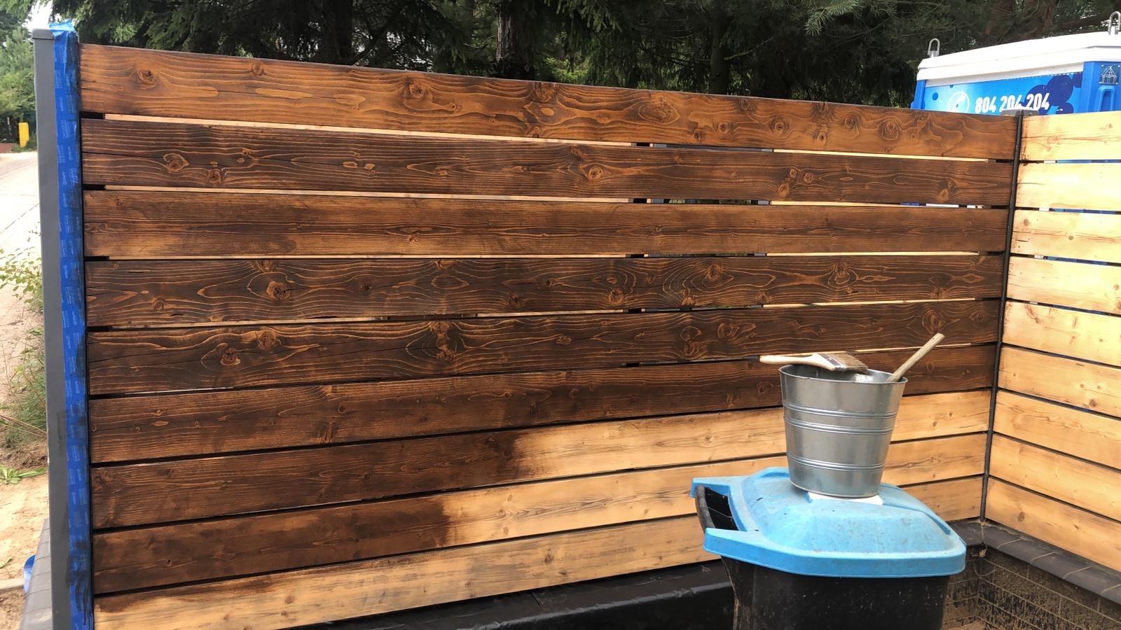 Częściowo pomalowane drewniane ogrodzenie poziome, z widocznym fragmentem surowego drewna i świeżo nałożoną bejcą, wiadro z pędzlem stoi na plastikowym koszu na śmieci.