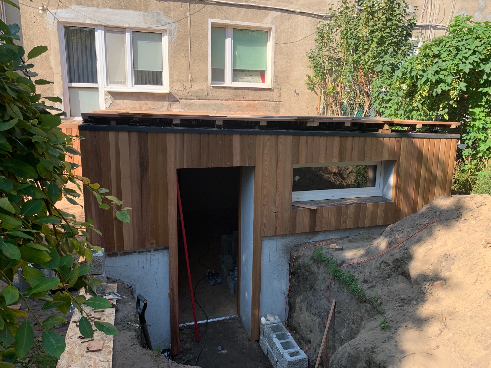 Budynek z drewnianą elewacją w trakcie budowy, widoczny wykop, częściowo odsłonięte fundamenty i okno. Na ścianie budynku widoczne dwa okna.