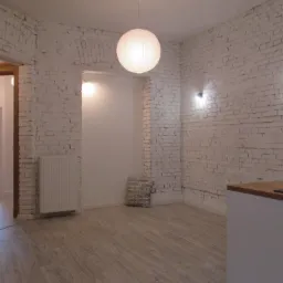 Jasne, przestronne wnętrze z białymi ceglanymi ścianami, drewnianą podłogą i papierową lampą wiszącą. Widoczne drzwi do kolejnego pomieszczenia, grzejnik pod ścianą i fragment blatu kuchennego...