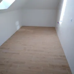 Ukończona podłoga wyłożona płytkami imitującymi drewno w jasnym, nowym pomieszczeniu na poddaszu z oknem dachowym i białymi ścianami.