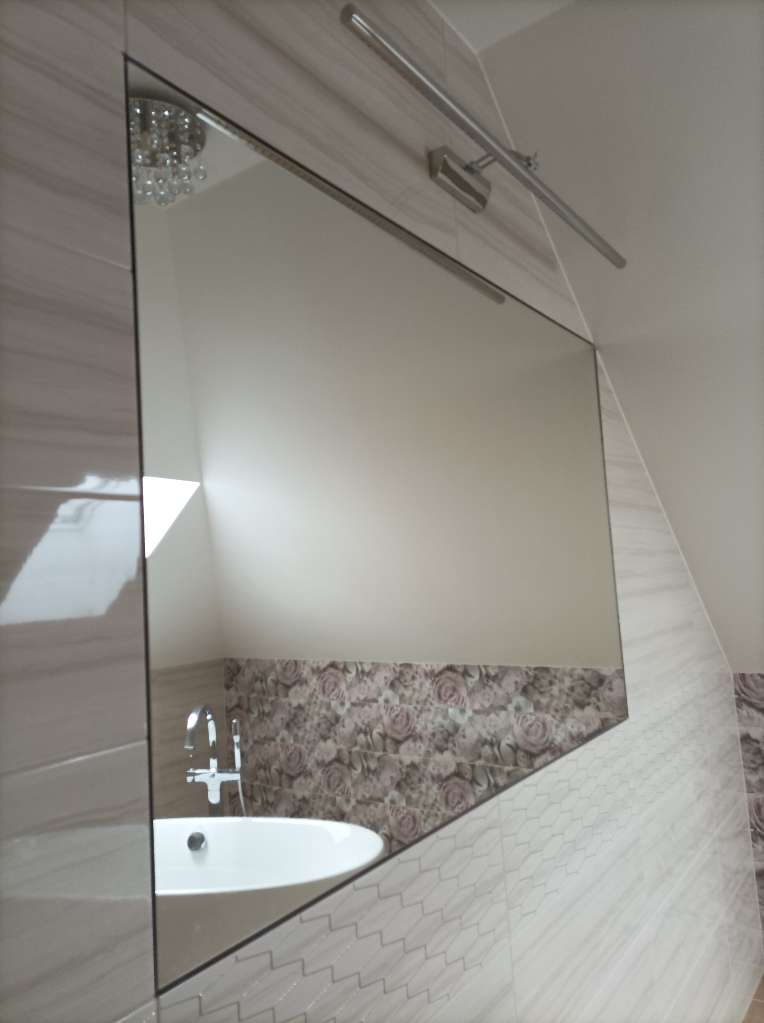 Nowoczesna łazienka z dużym lustrem, odbijającym kryształowy żyrandol, biała owalna umywalka z chromowaną baterią, ściany wyłożone płytkami w odcieniach beżu i szarości, dekoracyjny pas z motywem...