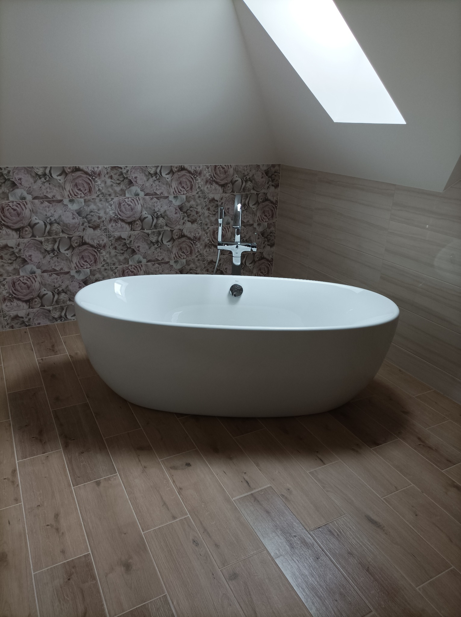 Wolnostojąca wanna owalna z chromowaną armaturą w łazience na poddaszu z oknem dachowym, wykończona płytkami imitującymi drewno na podłodze i ścianami w kwiatowe wzory.