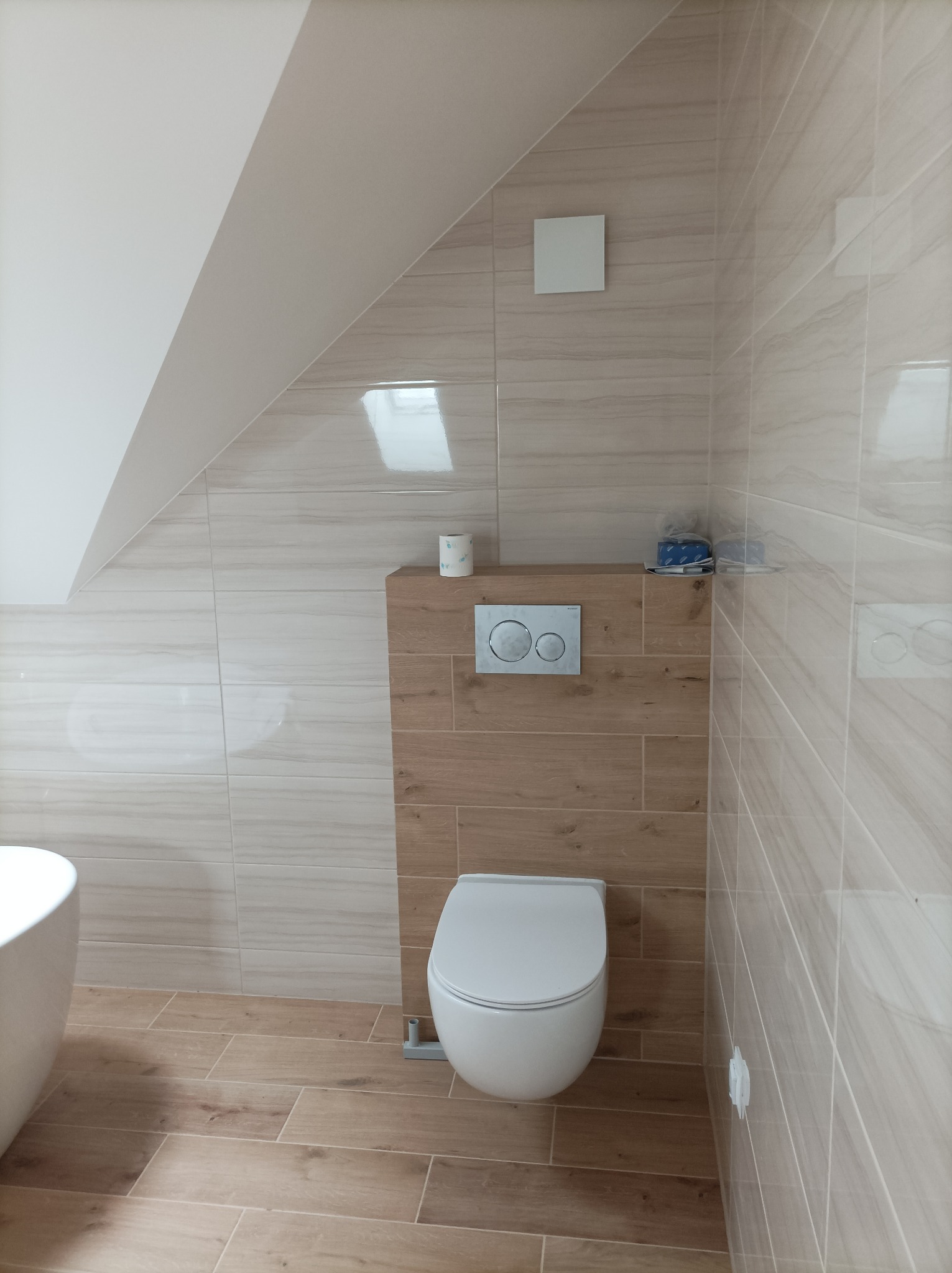 Nowoczesna toaleta podtynkowa z drewnopodobną zabudową w łazience na poddaszu, wykończona beżowymi płytkami imitującymi drewno, z widocznym fragmentem wanny.