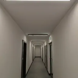 Długi korytarz biurowy z rzędem podłużnych, czarnych lamp LED zamontowanych na suficie, oświetlających szarymi płytkami wyłożoną podłogę i białe ściany z czarnymi futrynami drzwi.