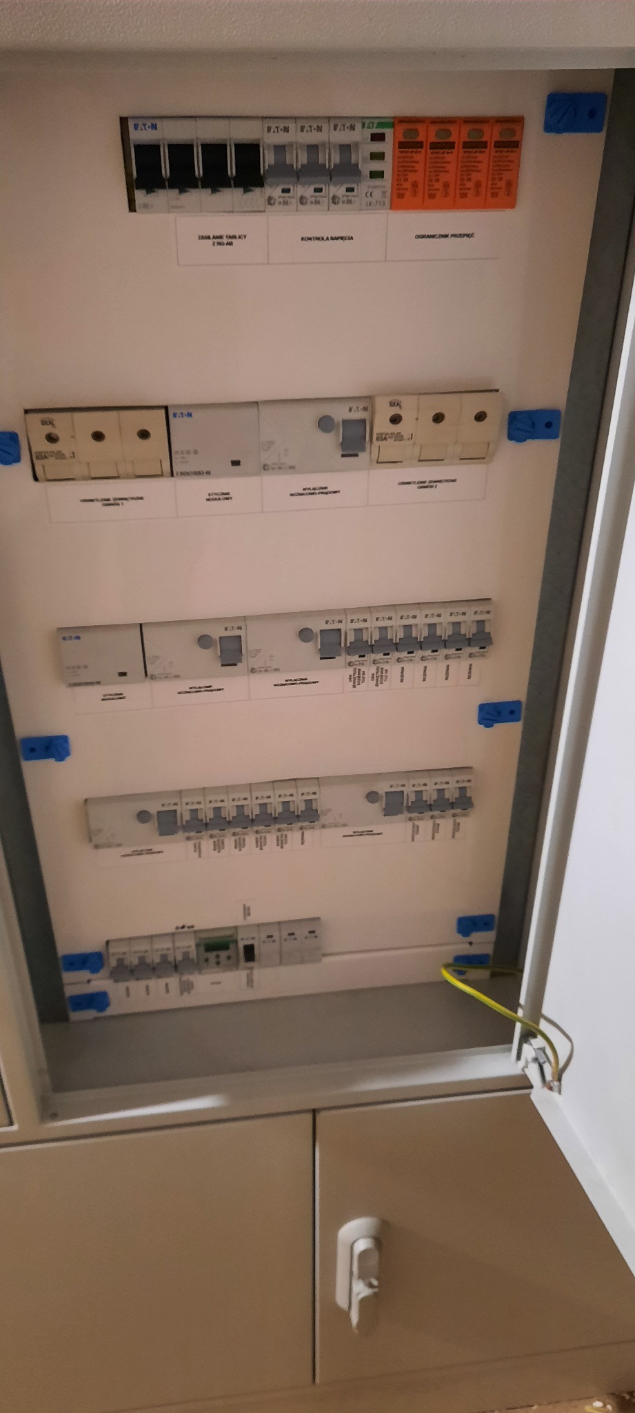 Otwarta szafa elektryczna z widocznymi bezpiecznikami, wyłącznikami różnicowoprądowymi i ogranicznikami przepięć marki Eaton, z żółto-zielonym przewodem uziemiającym.