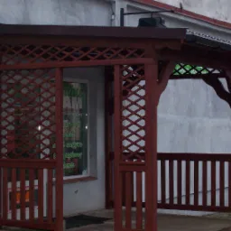 Ciemnobrązowa, drewniana pergola z kratownicą i daszkiem, przylegająca do budynku z oknem sklepowym, widoczna rynna.