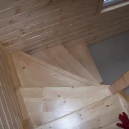 Wnętrze domu w trakcie wykańczania: drewniane schody spiralne z surowego drewna, widok z góry, z czerwoną rękawicą roboczą w rogu kadru.