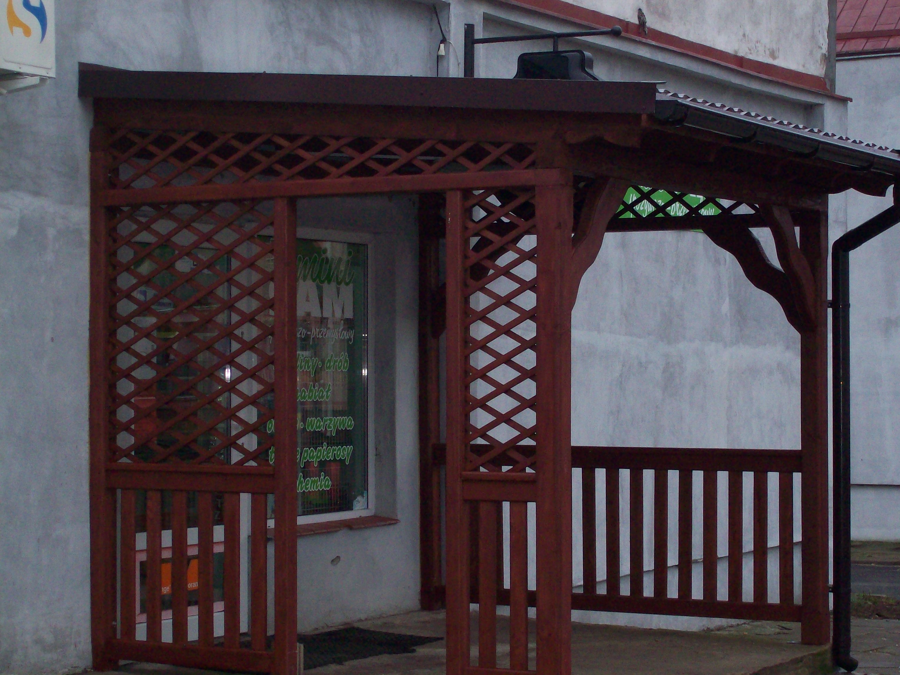 Ciemnobrązowa, drewniana pergola z kratownicą i daszkiem, przylegająca do budynku z oknem sklepowym, widoczna rynna.