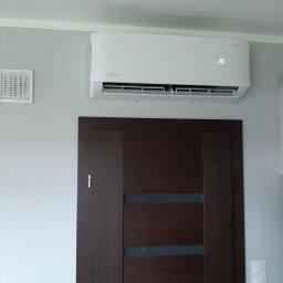 KLIMATIK - Biały klimatyzator Rotenso z wyświetlaczem ustawionym na 25 stopni Celsjusza, zamontowany nad brązowymi drzwiami w szarym pomieszczeniu z kratką wentylacyjną i włącznikiem światła.