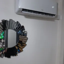 Biały klimatyzator Unical z wyświetlaczem ustawionym na 21 stopni Celsjusza, zamontowany na białej ścianie obok dekoracyjnego lustra słonecznego.