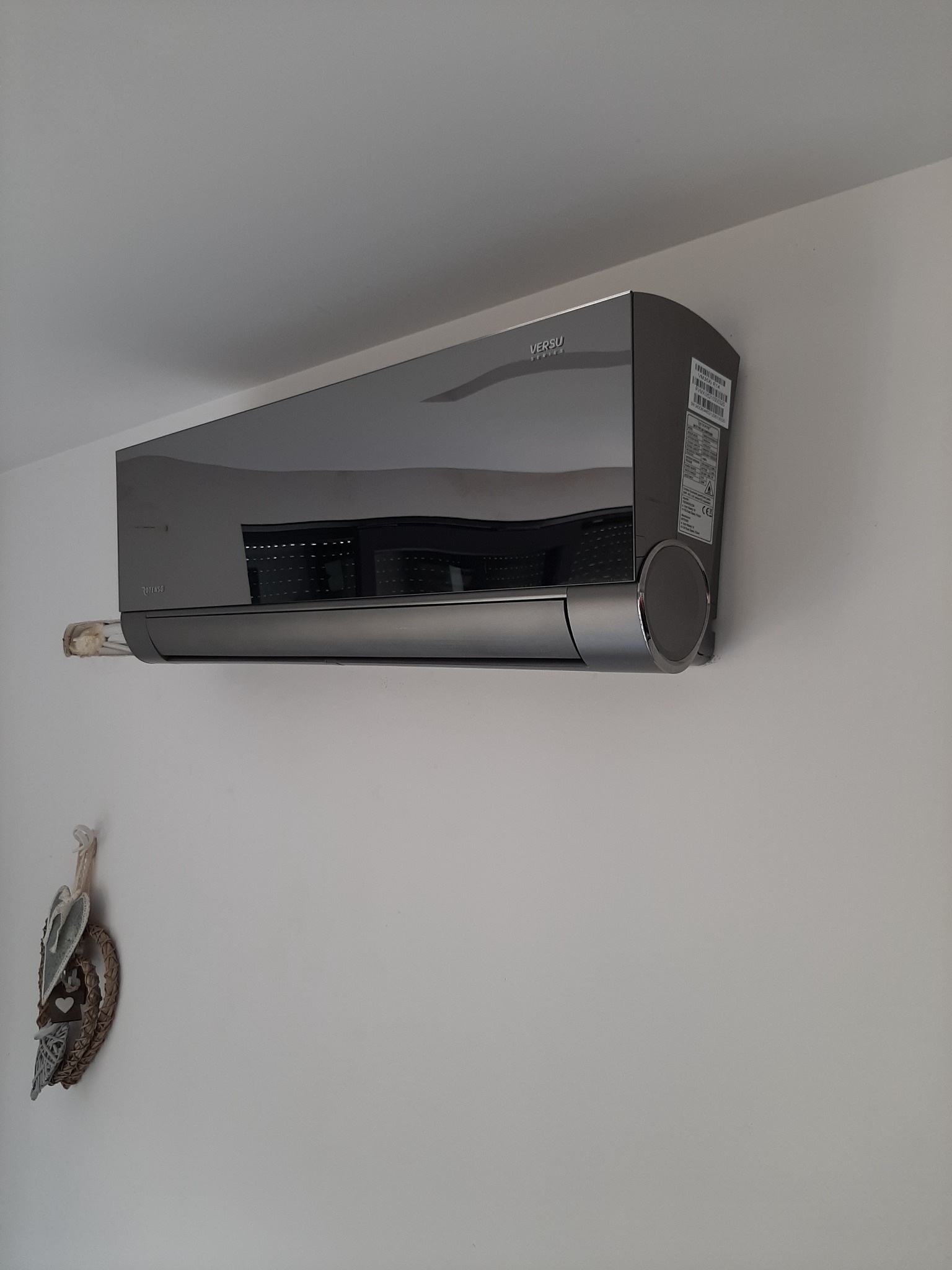 Szary klimatyzator marki Versu Series zamontowany na białej ścianie, z widocznym fragmentem dekoracji ściennej.