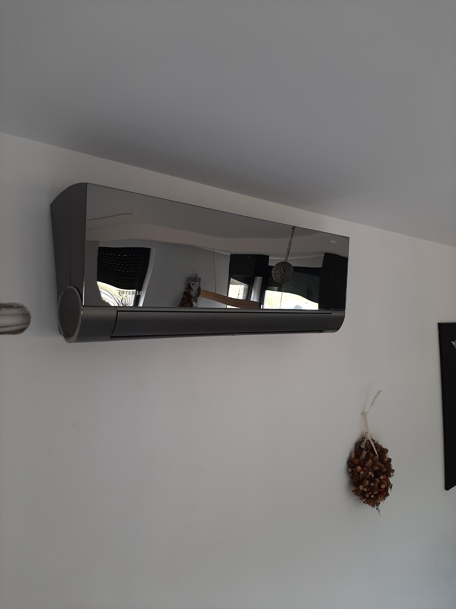 Nowoczesny, lustrzany klimatyzator ścienny w szarym kolorze, zamontowany w jasnym pomieszczeniu z widoczną dekoracją ścienną i odbiciem wnętrza w obudowie.