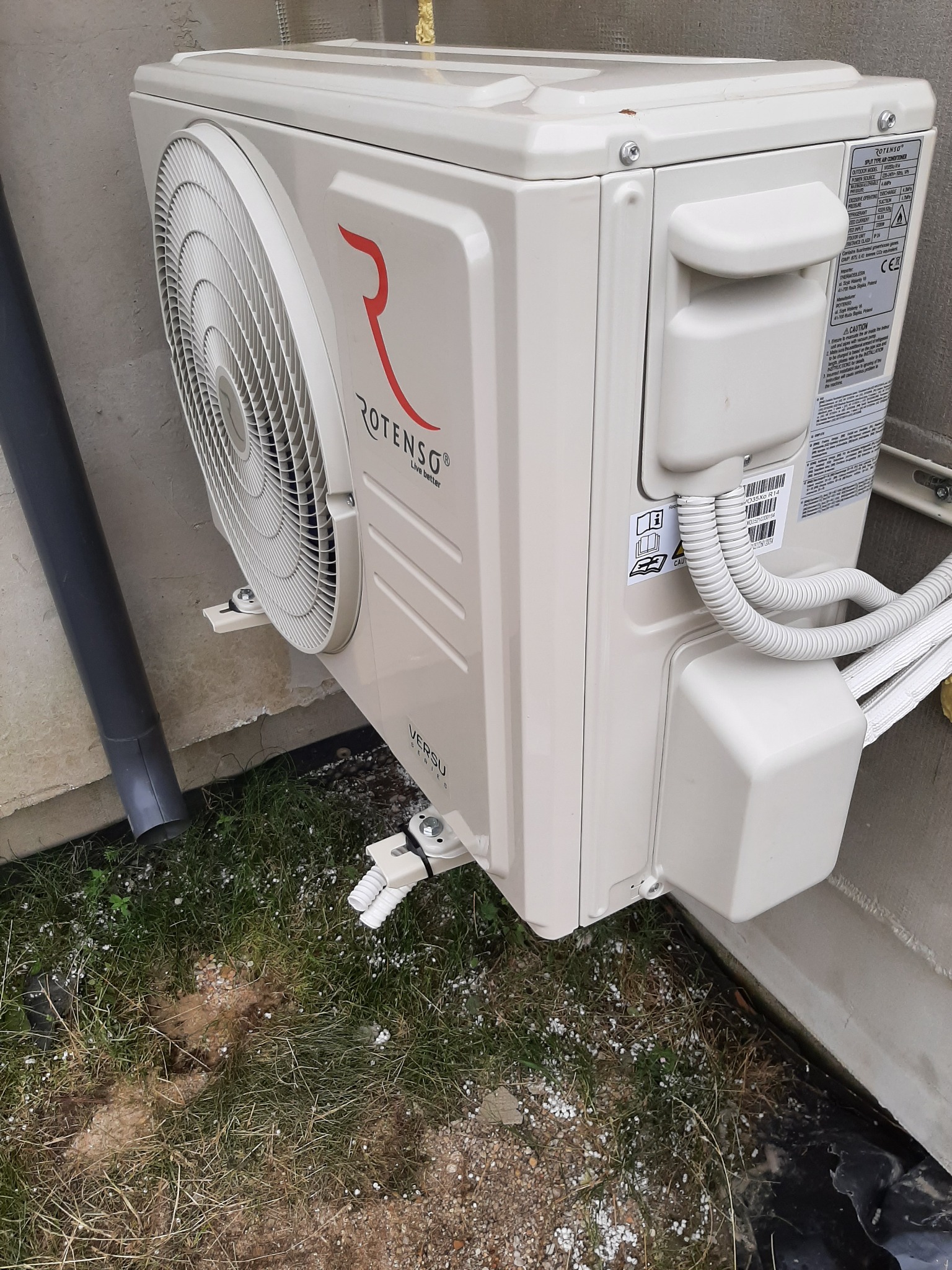 Zewnętrzna jednostka klimatyzacji Rotenso Versu zamontowana na ścianie budynku, widoczne przewody i rura odprowadzająca skropliny, trawa z drobnymi kamykami pod spodem.