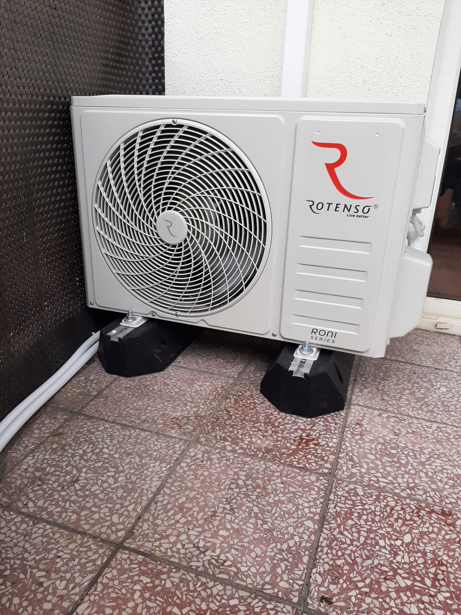 Zewnętrzna jednostka klimatyzatora Rotenso Roni Series, umieszczona na czarnych podkładkach antywibracyjnych na brązowej terakocie.