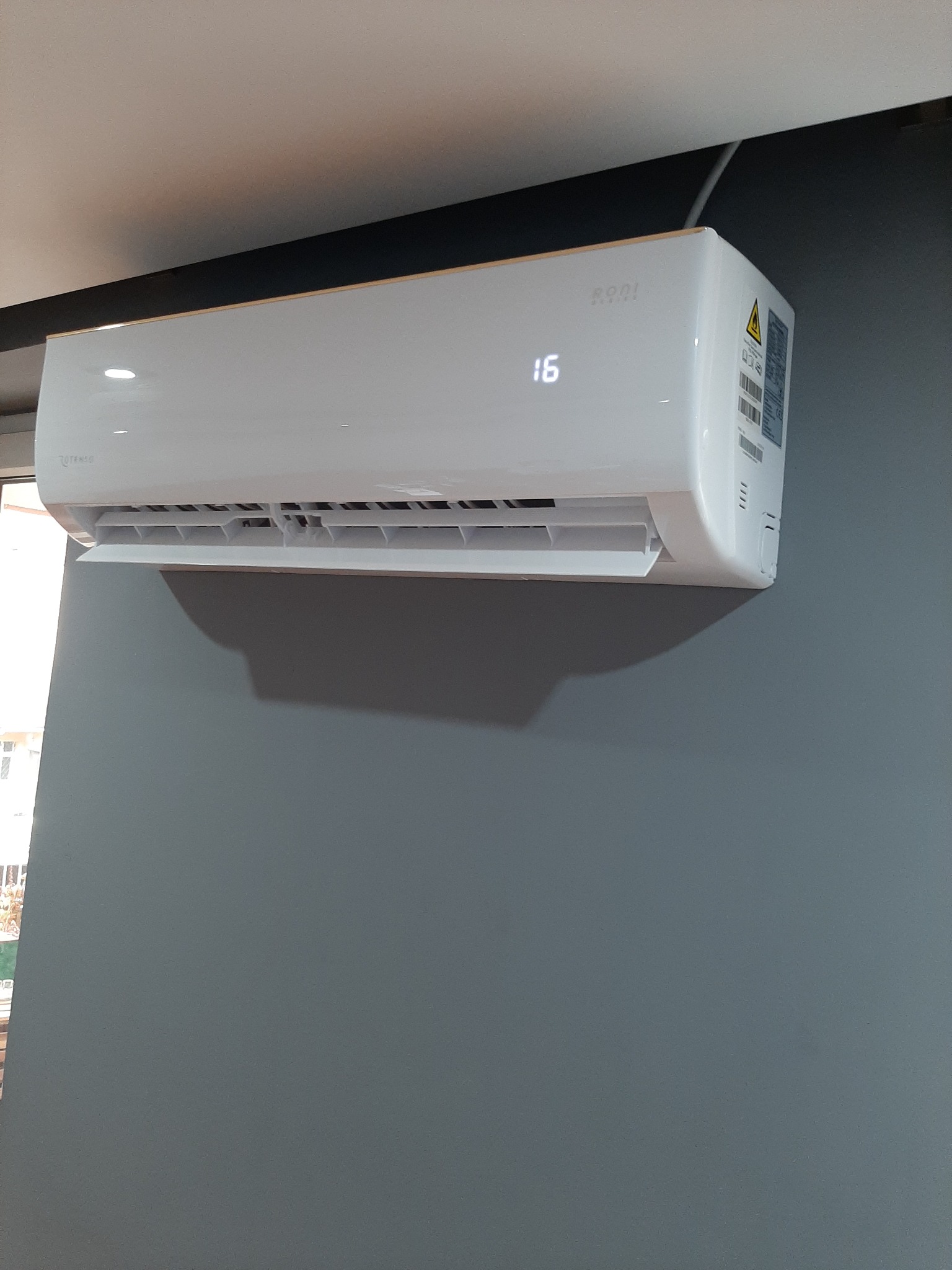 Biały klimatyzator marki Rooni zamontowany na szarej ścianie, wyświetlający temperaturę 16 stopni Celsjusza.