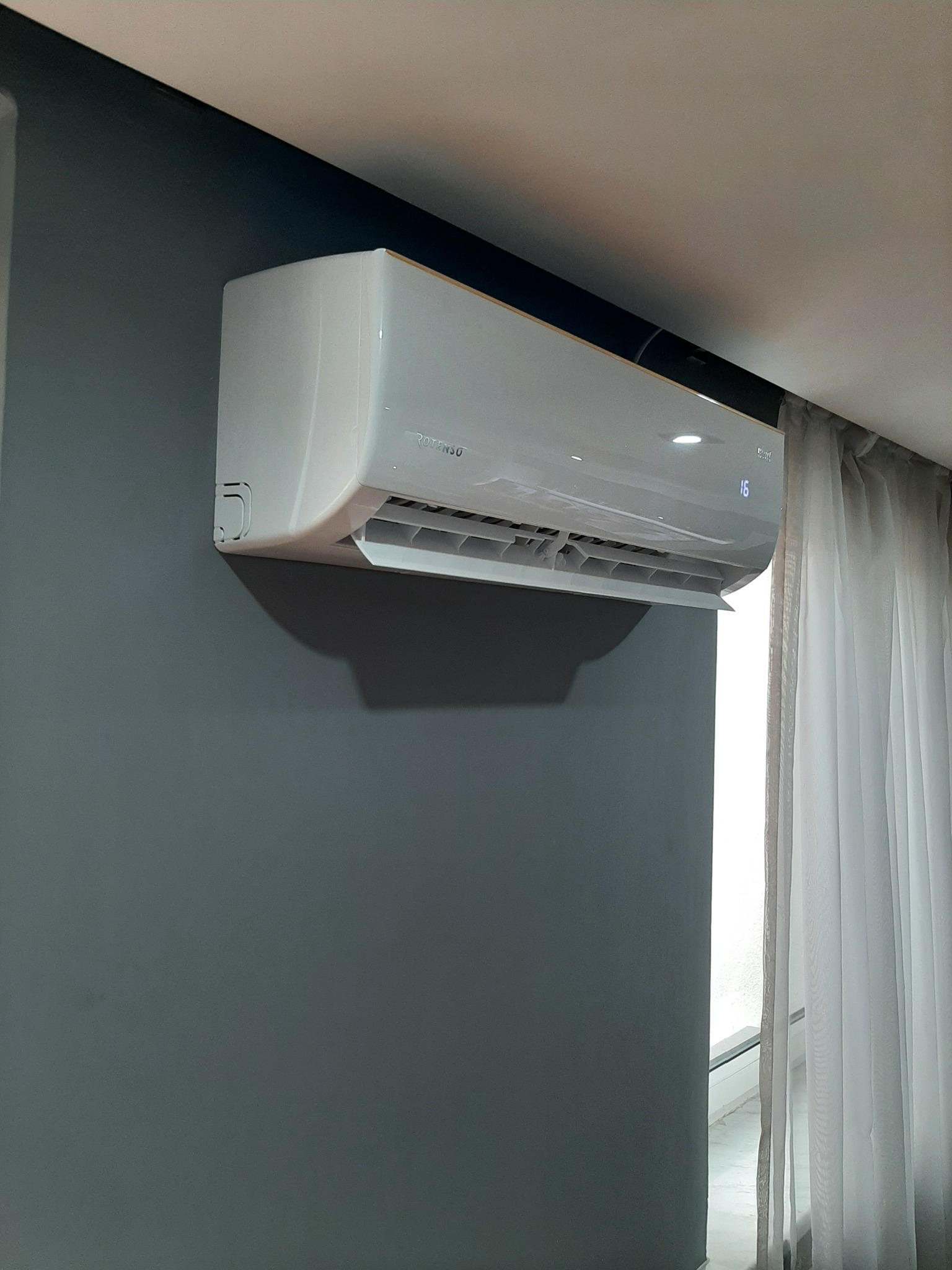 Biały klimatyzator marki Rotenso zamontowany na szarej ścianie w pomieszczeniu z zasłoniętym oknem.
