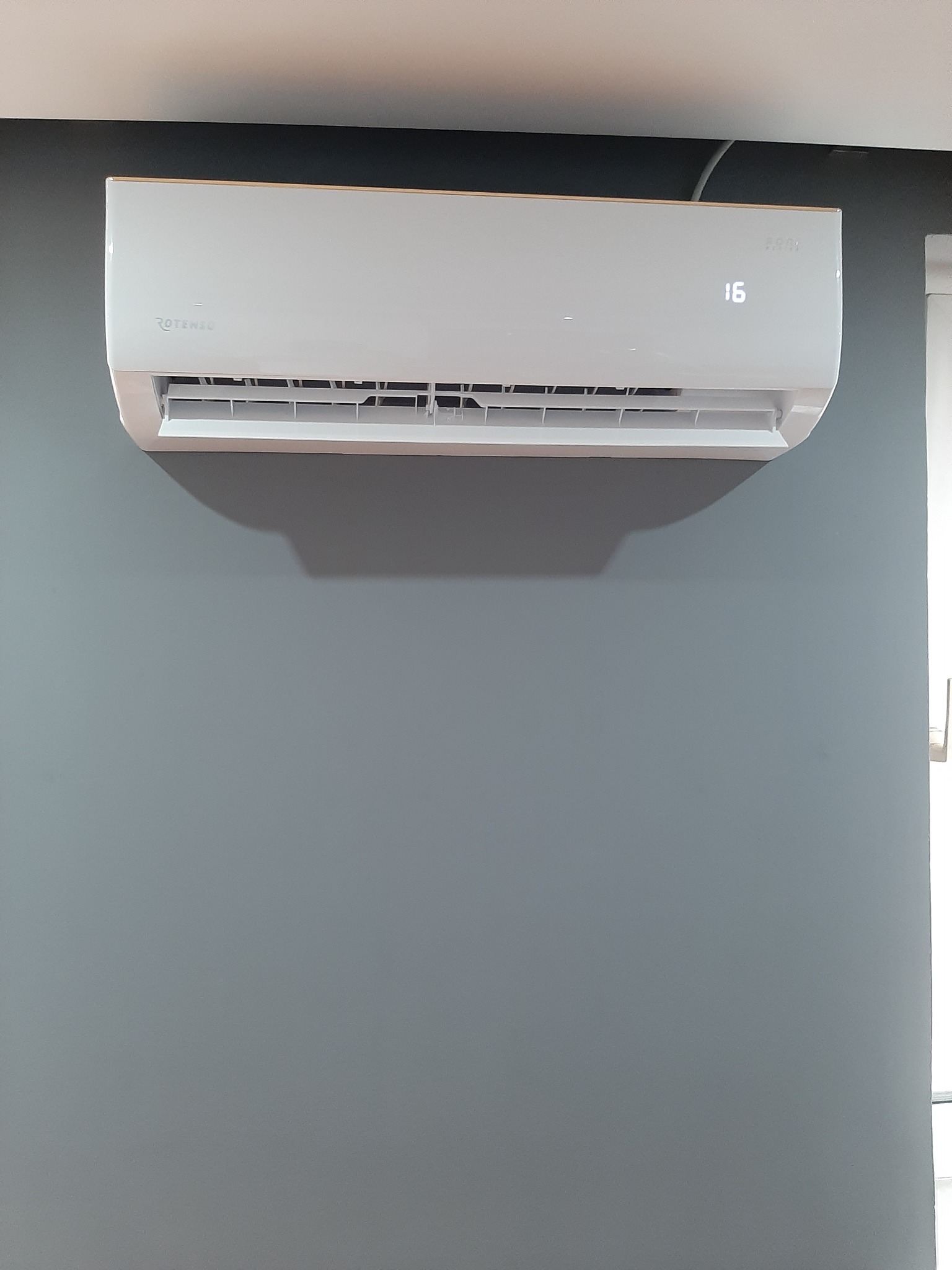 Biała klimatyzacja Rotenso zamontowana na szarej ścianie, wyświetlająca temperaturę 16 stopni Celsjusza.