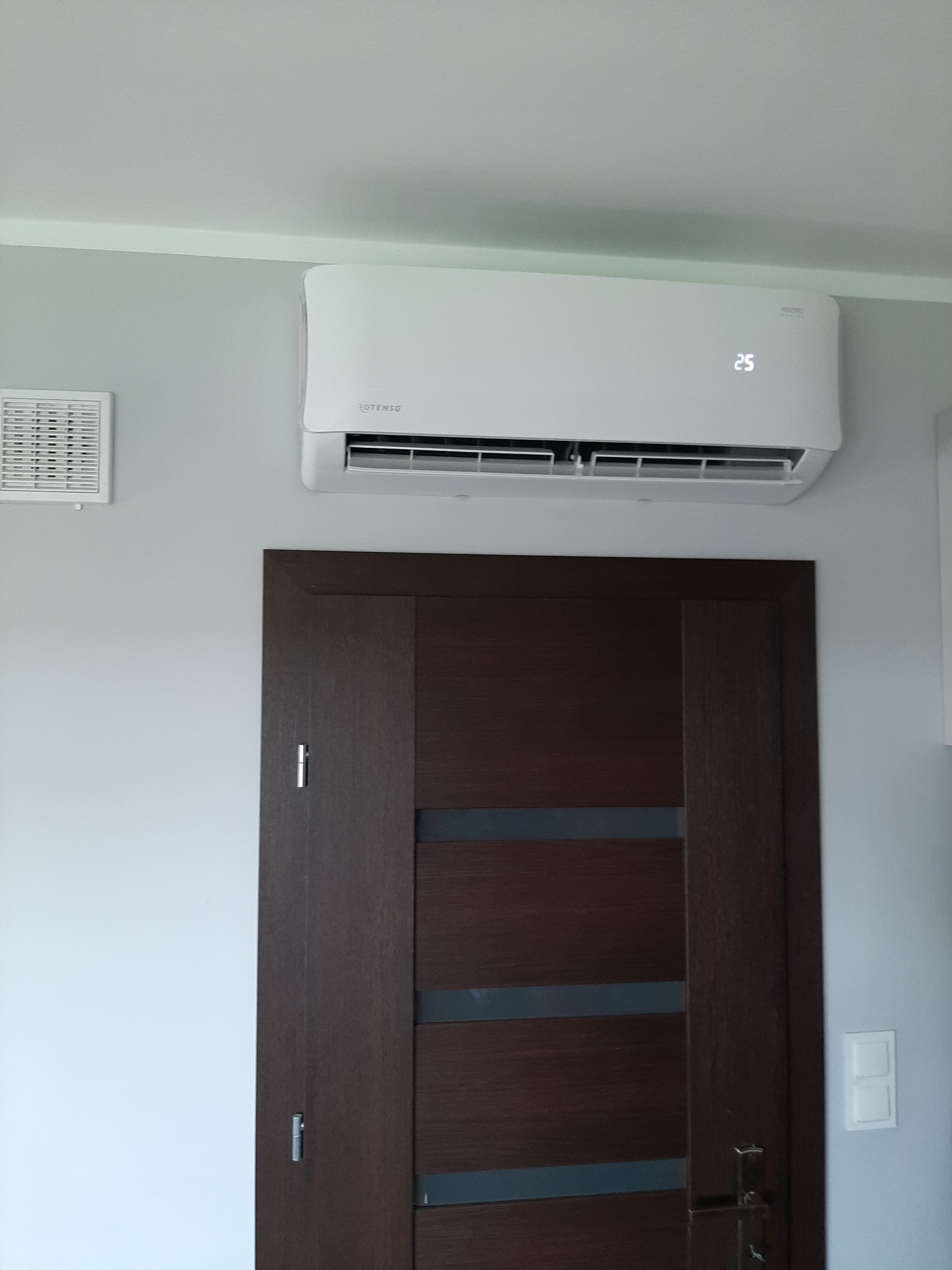 Biały klimatyzator Rotenso z wyświetlaczem ustawionym na 25 stopni Celsjusza, zamontowany nad brązowymi drzwiami w szarym pomieszczeniu z kratką wentylacyjną i włącznikiem światła.