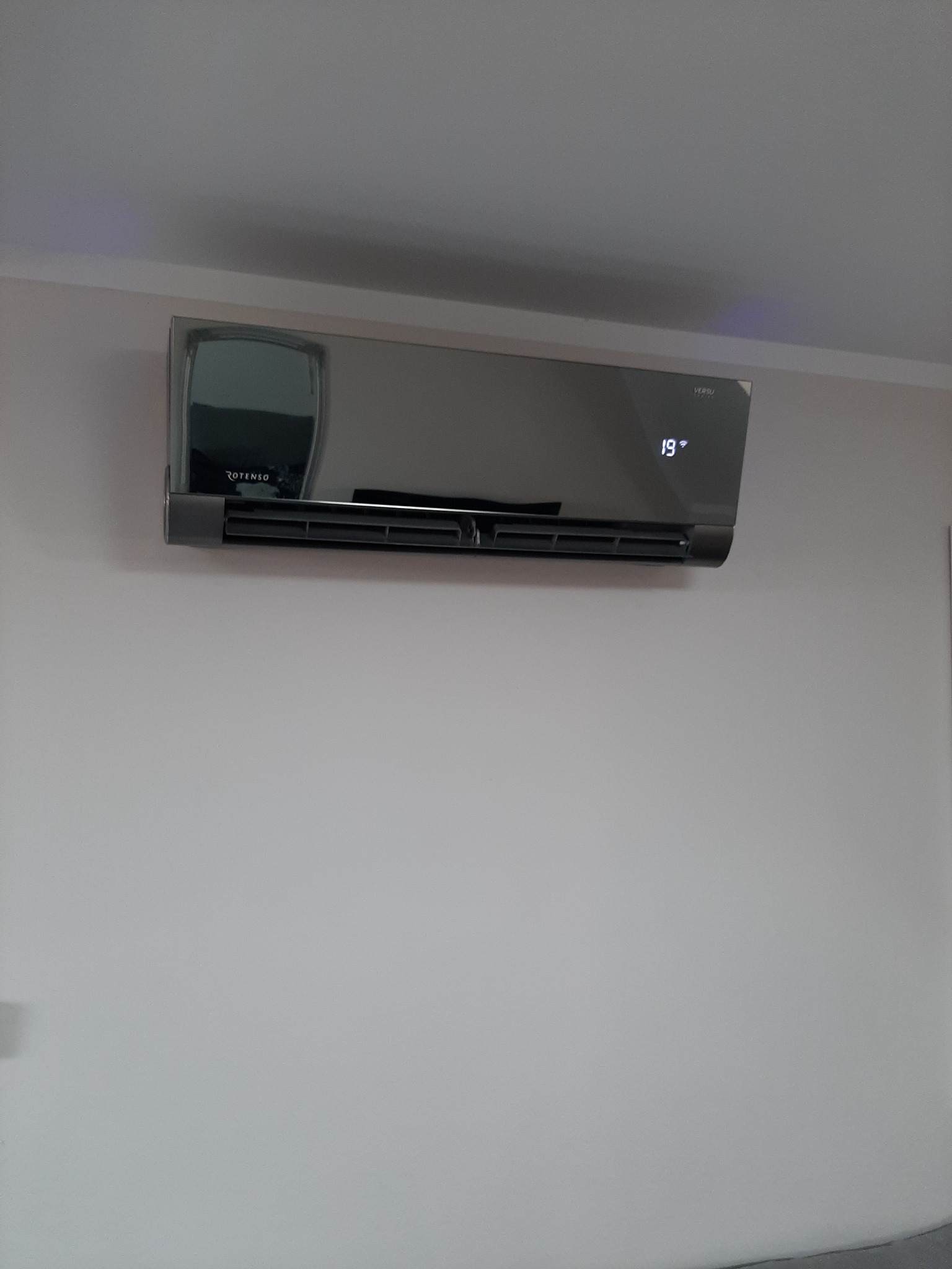 Czarny klimatyzator marki Rotenso zamontowany na białej ścianie, wyświetlający temperaturę 19 stopni Celsjusza.