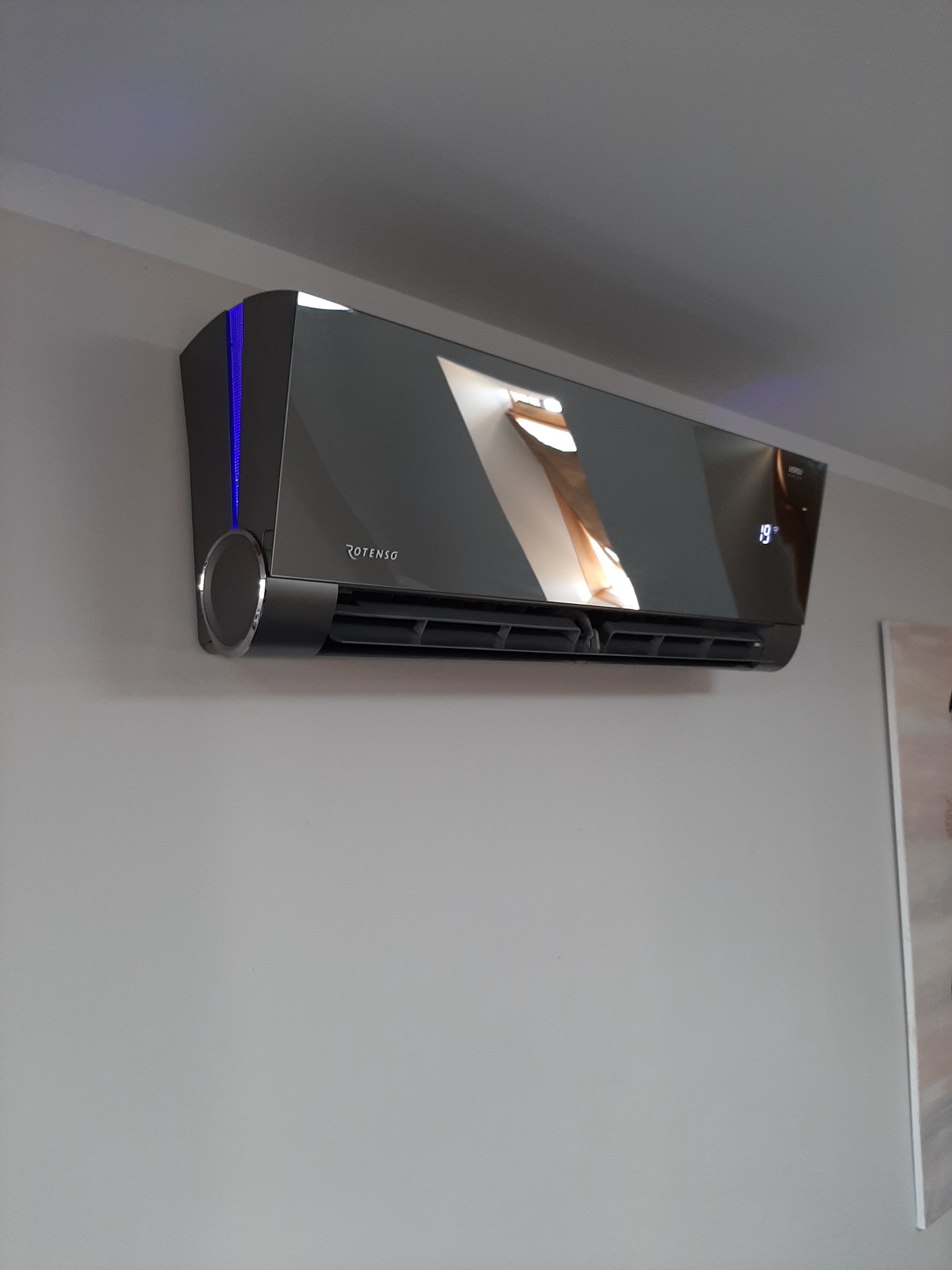 Nowoczesny klimatyzator Rotenso w ciemnej obudowie z wyświetlaczem temperatury 19 stopni Celsjusza, zamontowany na jasnej ścianie w pomieszczeniu.