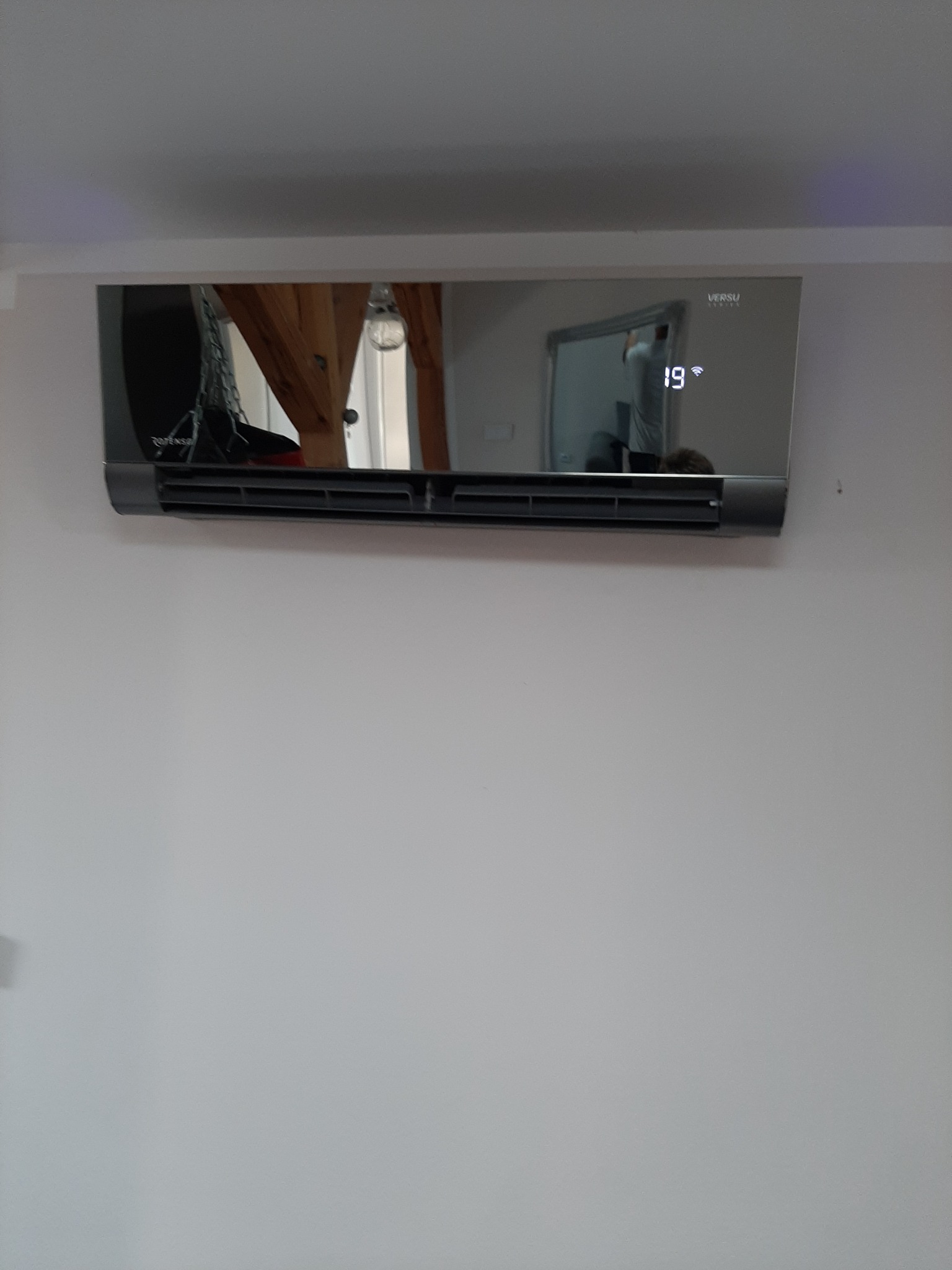 Szary klimatyzator Versu zamontowany na białej ścianie, wyświetlający temperaturę 19 stopni, z widocznym odbiciem wnętrza pokoju.