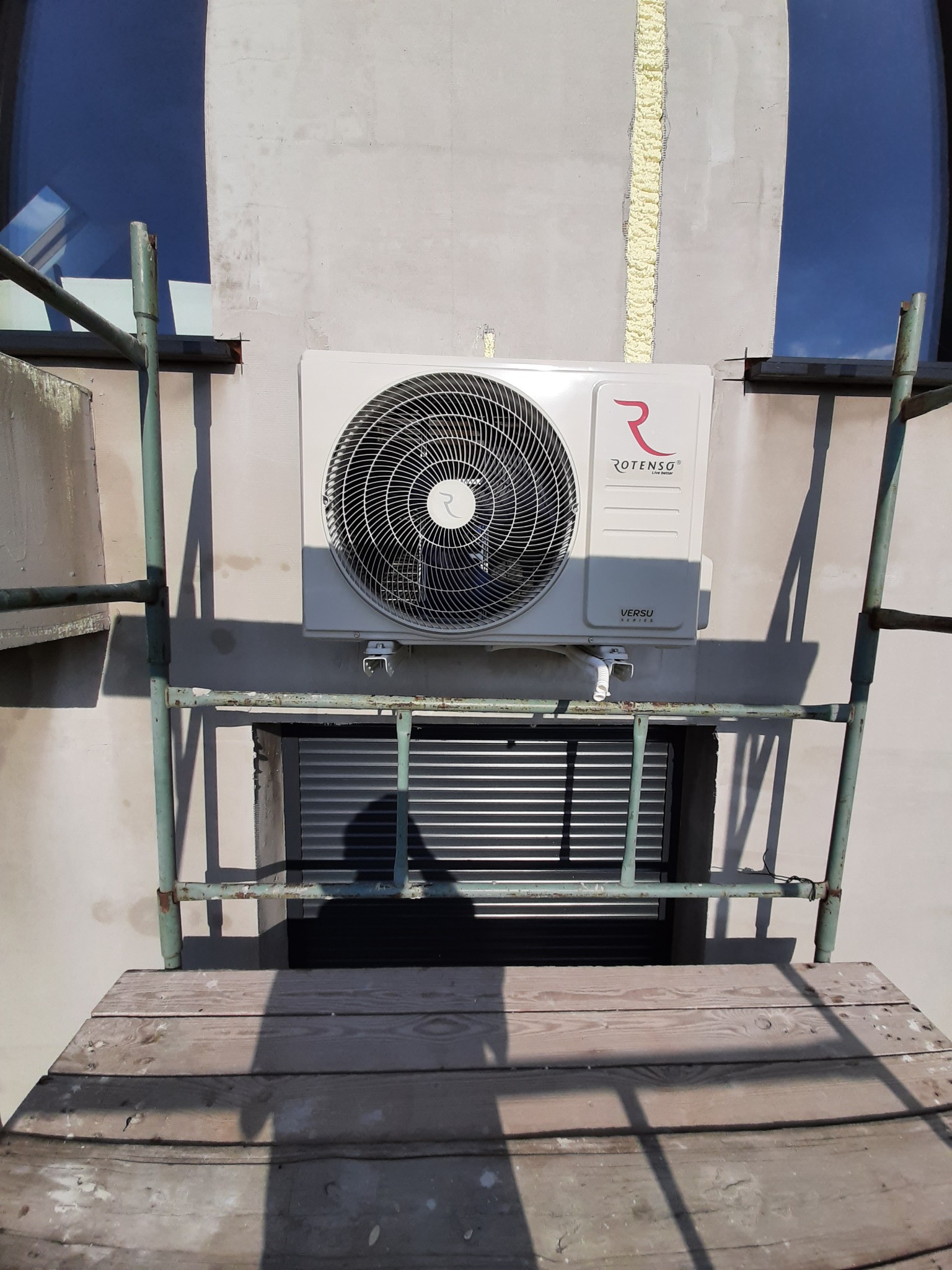 Zamontowany zewnętrzny moduł klimatyzatora Rotenso Versu Series na szarej ścianie budynku, widoczny z poziomu rusztowania, z pianką montażową uszczelniającą szczelinę obok.