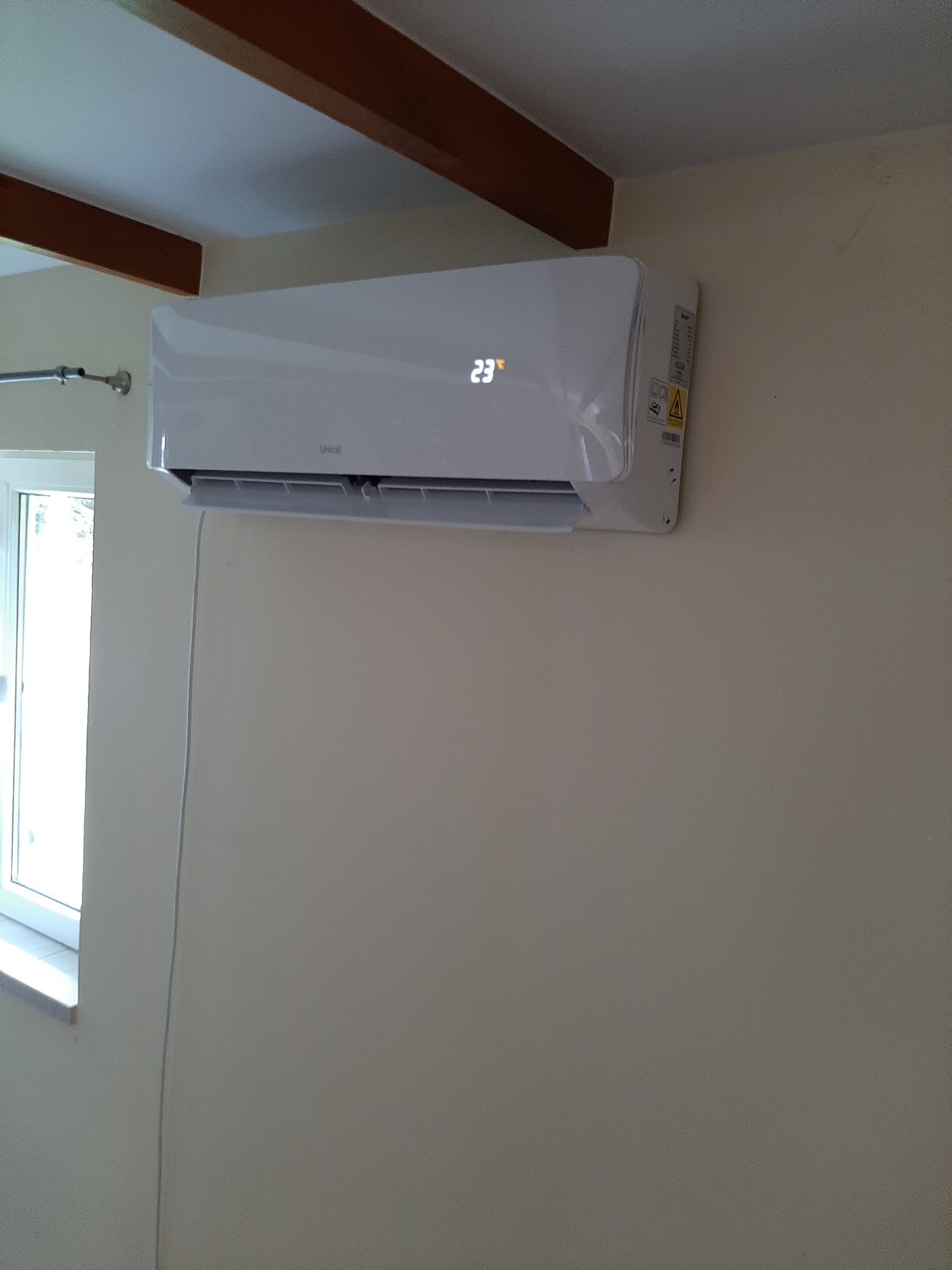 Biały klimatyzator ścienny z wyświetlaczem temperatury 23 stopnie Celsjusza, zamontowany w pokoju z beżowymi ścianami i drewnianymi belkami stropowymi, widoczny fragment okna.