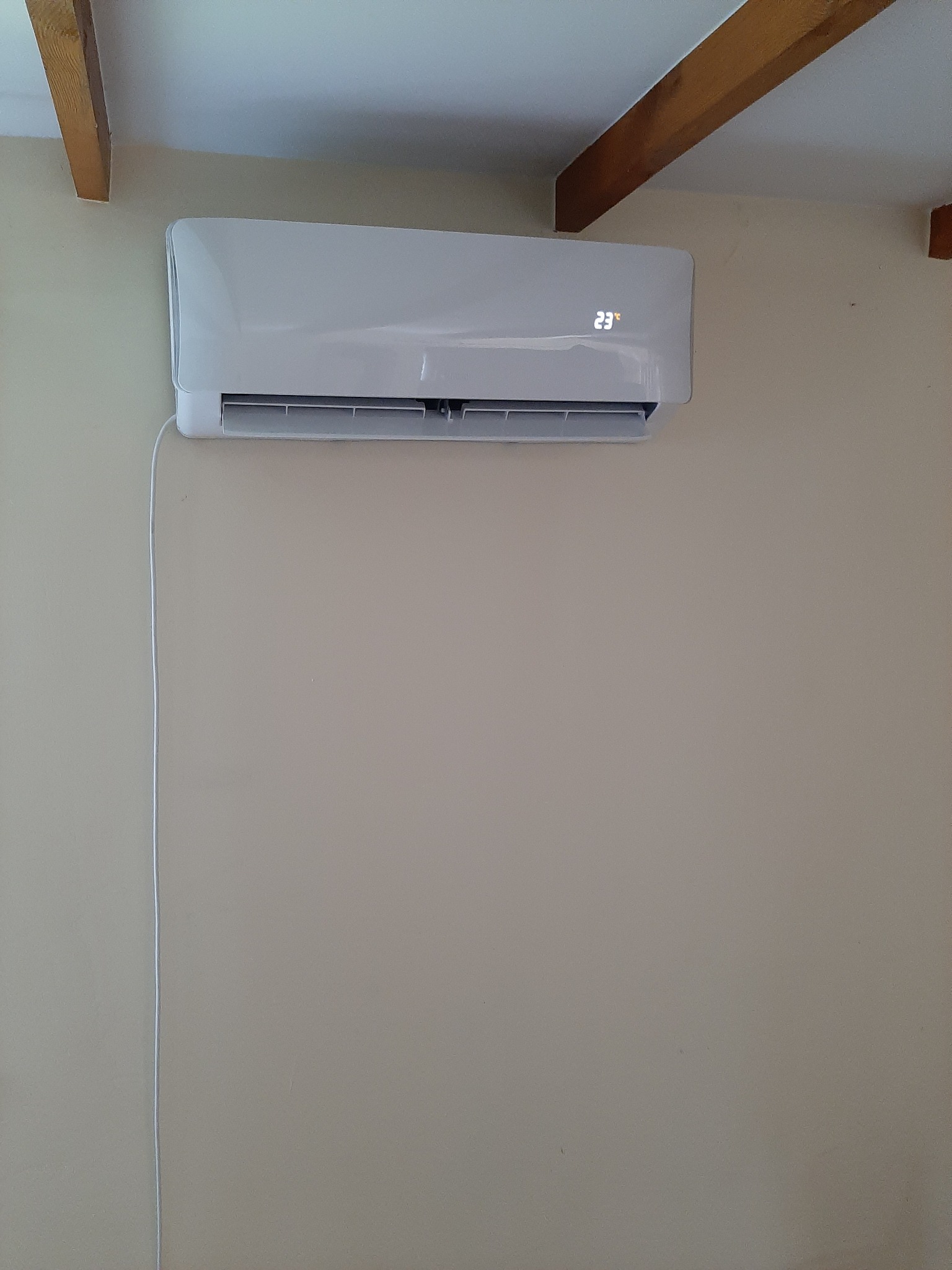 Biały klimatyzator ścienny z wyświetlaną temperaturą 23 stopnie Celsjusza zamontowany w pokoju z beżowymi ścianami i drewnianymi belkami stropowymi.