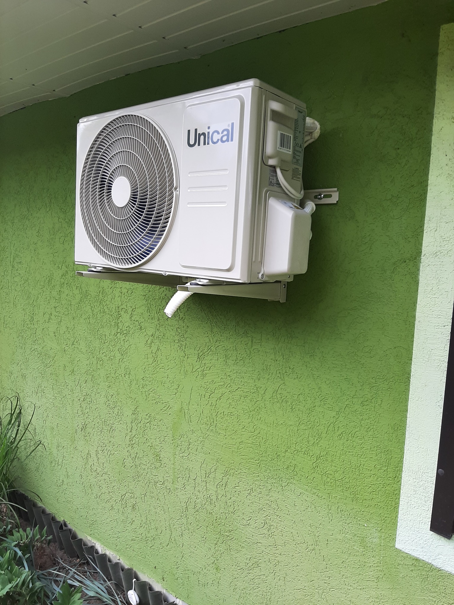 Zamontowana na zielonej, teksturowanej ścianie zewnętrzna jednostka klimatyzacji marki Unical z widoczną rurką odprowadzającą skropliny.