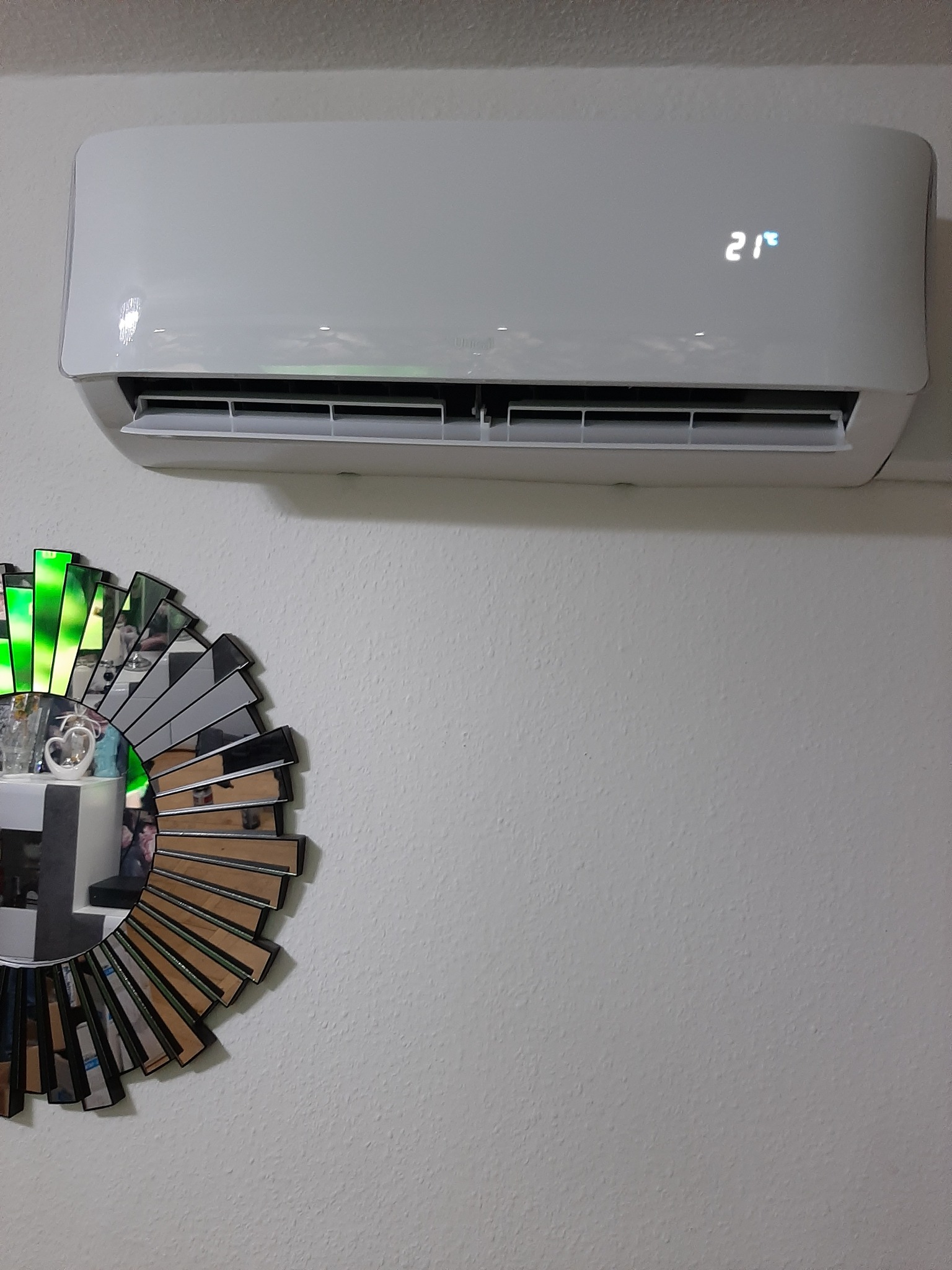 Biały klimatyzator ścienny z wyświetlaczem ustawionym na 21 stopni, zamontowany nad dekoracyjnym lustrem w kształcie słońca z elementami zieleni.