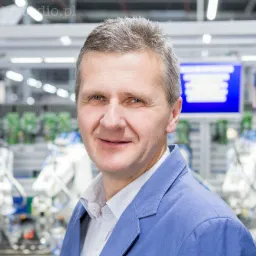 Portret uśmiechniętego mężczyzny w niebieskiej marynarce na tle zautomatyzowanej linii produkcyjnej z robotami i ekranem LCD.